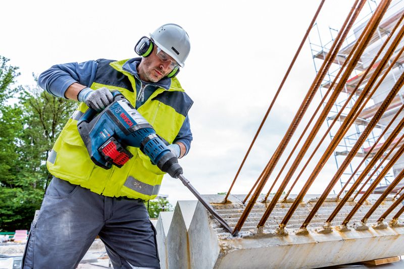 A person wearing safety equipment drills into concrete with a large cordless rotary hammer.