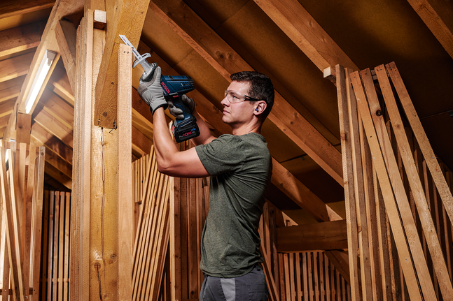 Person wearing safety equipment cuts wood in an attic using a cordless reciprocating saw.