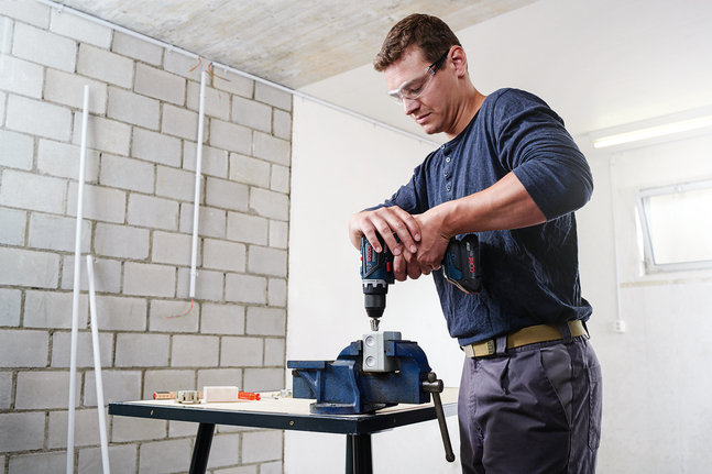 Person wearing safety equipment drills into a metal piece secured in a vise.
