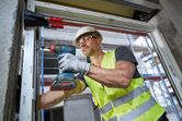 A person wearing safety equipment drives a screw into a window frame with a cordless drill.