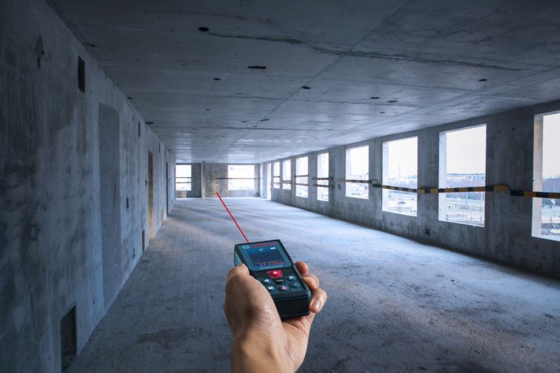 Person using a laser measure to determine distance in an unfinished concrete room.