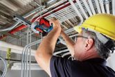 A person wearing safety equipment uses a cordless band saw to cut metal conduit overhead.