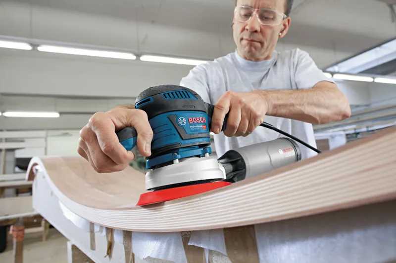 A person wearing safety equipment sands a curved wooden surface with a power tool.