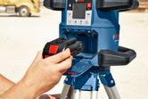 Person inserting a battery into a laser leveling tool on a tripod outdoors.