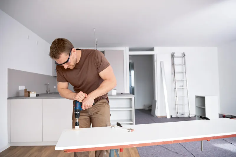 A person wearing safety equipment drills into a white board in a modern kitchen.