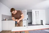 A person wearing safety equipment drills into a white board in a modern kitchen.