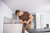 A person wearing safety equipment uses a cordless combi drill on a kitchen cabinet.