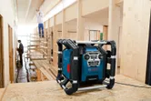 Jobsite radio sitting on plywood, with two people working on scaffolding in a wooden building.