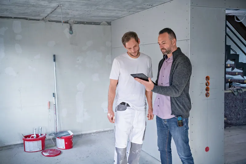 Two people review construction plans on a tablet, one holds a digital measuring device.