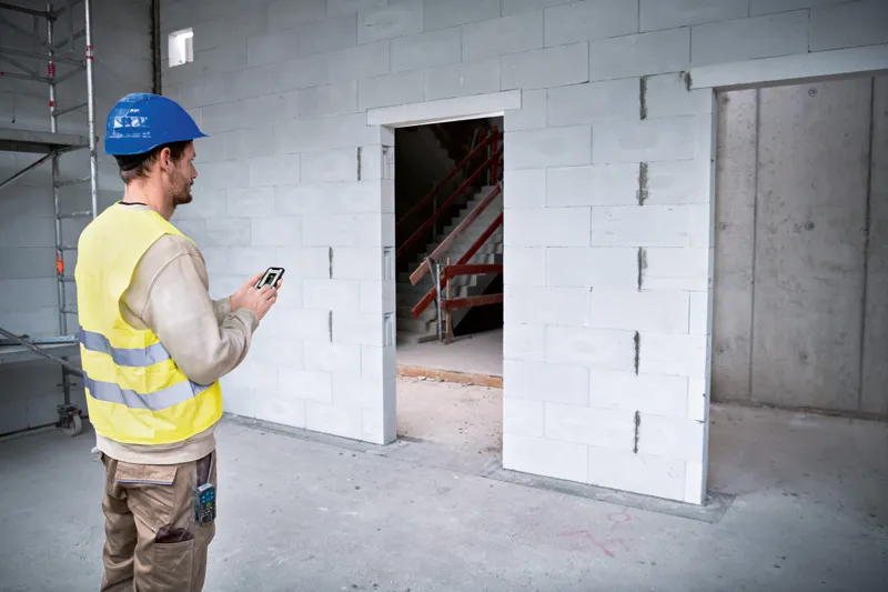 GLM 50-27 CG Person wearing safety equipment uses a laser measure to check doorway dimensions on a construction site.