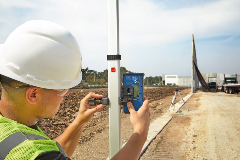 A person wearing safety equipment adjusts a laser receiver on a leveling rod at a construction site.