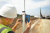 A person wearing safety equipment adjusts a laser receiver on a leveling rod at a construction site.