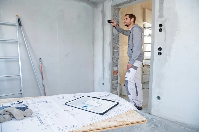 GLM 50-27 CG A person uses a laser measure to check wall distance at a construction site.