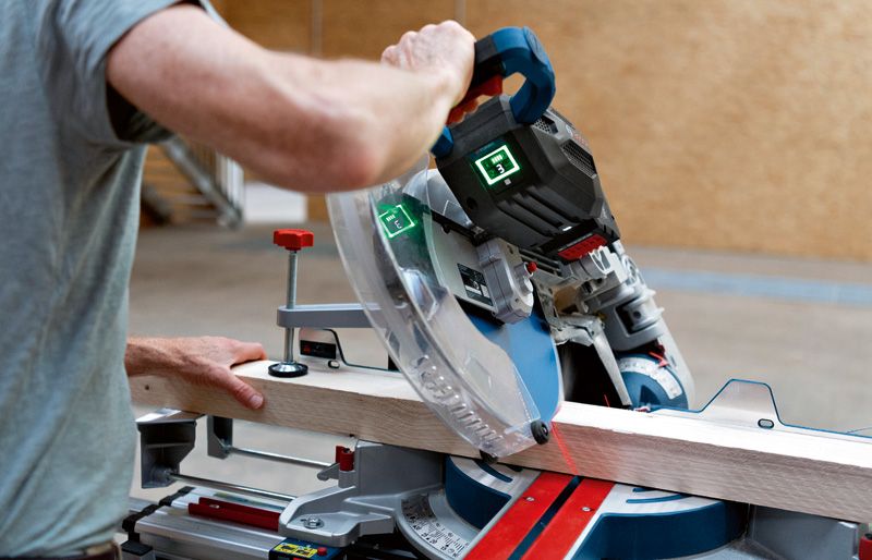 A person operates a cordless mitre saw to cut a wooden board indoors.