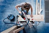 A person wearing safety equipment uses a cordless mitre saw to cut a wooden plank.