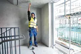 A person wearing safety equipment uses a point laser to mark the ceiling in a concrete room.