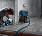 A person wearing safety equipment grinds concrete with a dust extractor connected by hose.