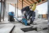 A person wearing safety equipment uses a wet/dry extractor and cutting tool on a concrete floor.