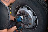 A person wearing safety equipment uses an impact wrench to tighten bolts on a truck wheel.