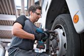 A person wearing safety equipment uses a cordless impact wrench on a truck wheel.
