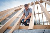 A person wearing safety equipment uses a cordless impact wrench to fasten wood beams.