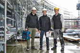 Three workers wearing safety equipment stand at a construction site, one holding a power tool.