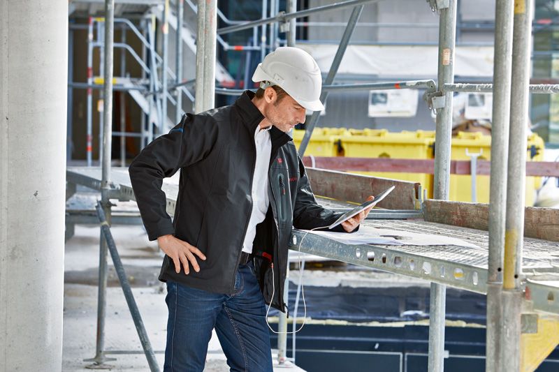 A person wearing safety equipment and a heated jacket inspects a tablet at a construction site.