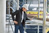 A person wearing safety equipment and a heated jacket inspects a tablet at a construction site.