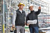 Two workers wearing safety equipment discuss building plans at a construction site.