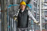 A person wearing safety equipment and heated vest carries a metal beam at a construction site.