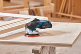 Cordless orbital sander resting on a wooden board in a carpentry workshop.