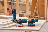 Three electric sanders rest on a wooden workbench in a carpentry workshop.