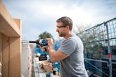A person wearing safety equipment uses a cordless impact wrench to drive a bolt into wood.