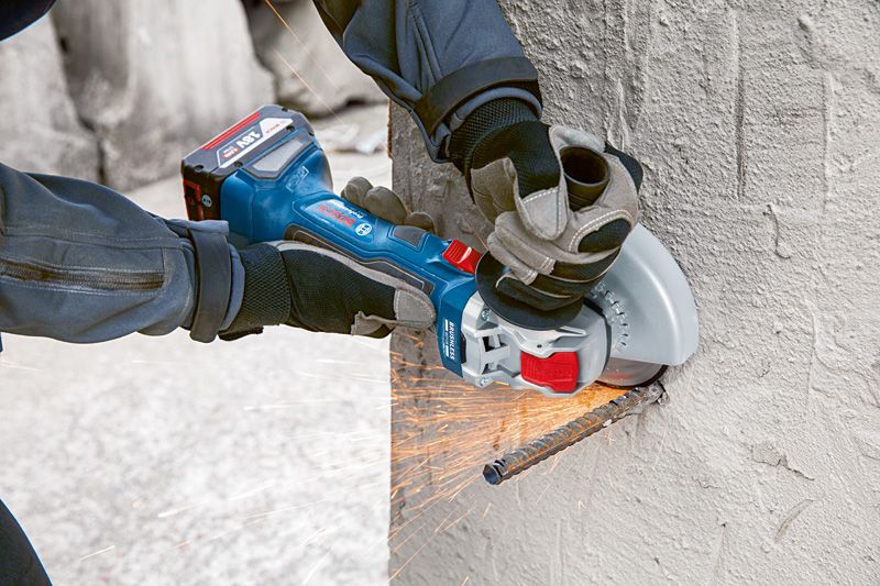 A person wearing safety equipment cuts a concrete wall with a cordless angle grinder.