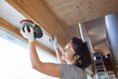 A person wearing safety equipment uses a sander to smooth a wooden beam indoors.