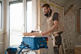A person wearing safety equipment cuts wood using a cordless table saw in a room under renovation.