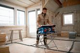 A person wearing safety equipment sets up a cordless table saw in a room under renovation.