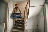 A person carrying a cordless table saw down a narrow staircase at a construction site.