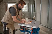 A person wearing safety equipment cuts wood on a cordless table saw in a construction area.