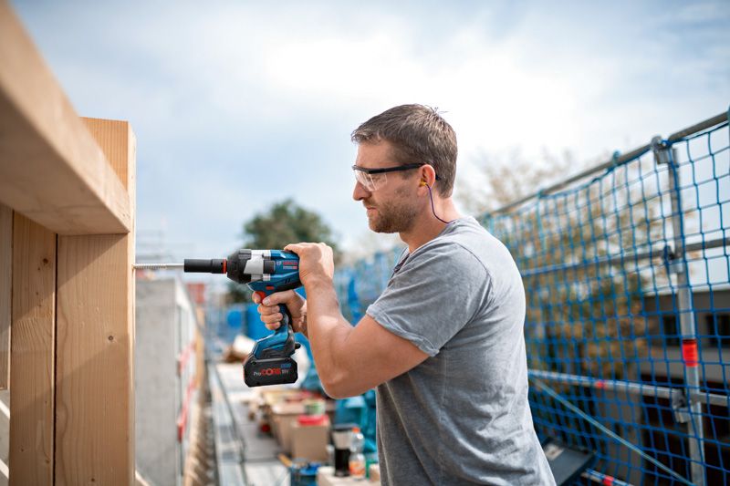 A person wearing safety equipment uses a cordless impact wrench to drive a screw into wood.