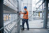 A person wearing safety equipment uses a cordless impact wrench on a steel beam.
