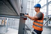 A person wearing safety equipment uses a cordless impact wrench on a steel beam.