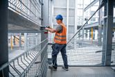 A person wearing safety equipment uses a cordless impact wrench on a steel beam.