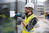 A person wearing safety equipment uses a cordless heat gun on a glass panel.