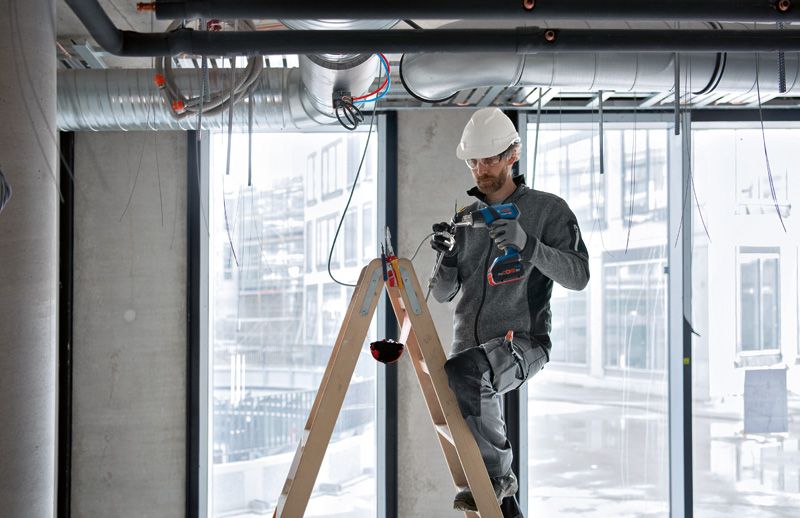 GHG 18V-50 A person wearing safety equipment uses a cordless heat gun on a ladder near ceiling ducts.