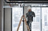 A person wearing safety equipment uses a cordless heat gun on a ladder near ceiling ducts.