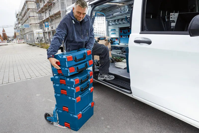 A person wearing safety equipment loads stacked toolboxes onto a dolly beside a work van.
