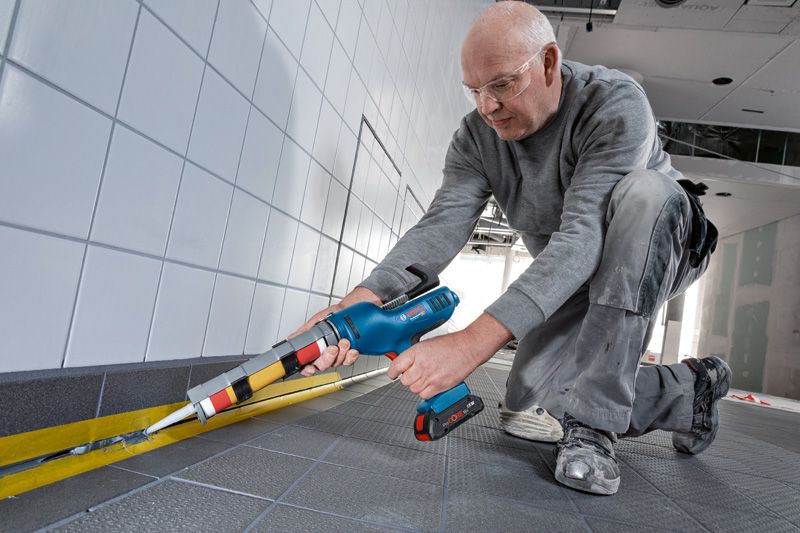 A person wearing safety equipment applies sealant to floor tiles using a power caulking gun.