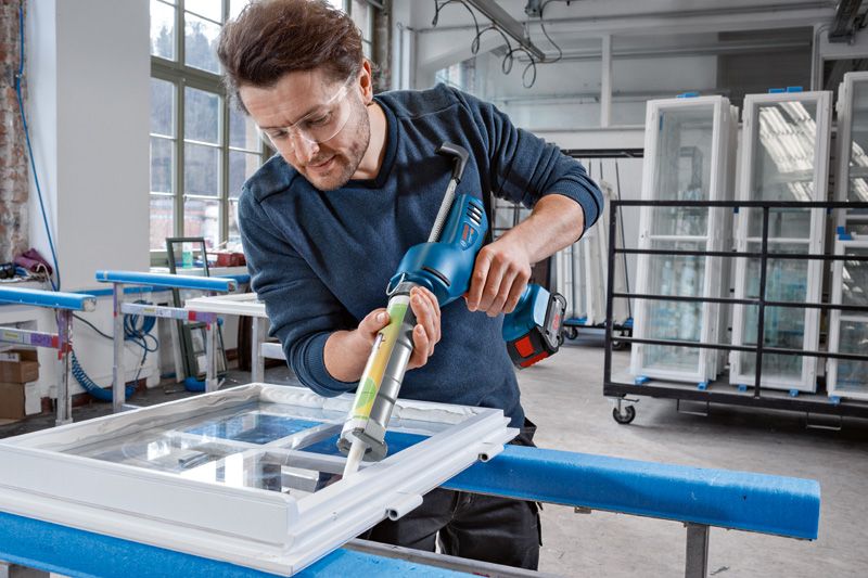 A person wearing safety equipment applies sealant to a window frame using a cordless caulking gun.