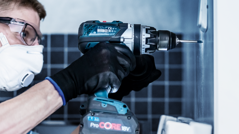 Person wearing safety equipment uses a cordless drill to drive a screw into a wall.